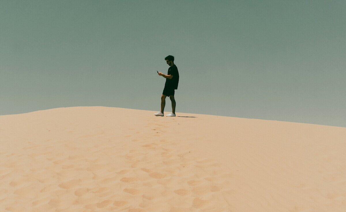 Mann steht auf einer Düne und guckt auf das Smartphone © Bryan Williams / Unsplash