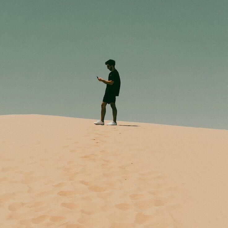 Mann steht auf einer Düne und guckt auf das Smartphone © Bryan Williams / Unsplash